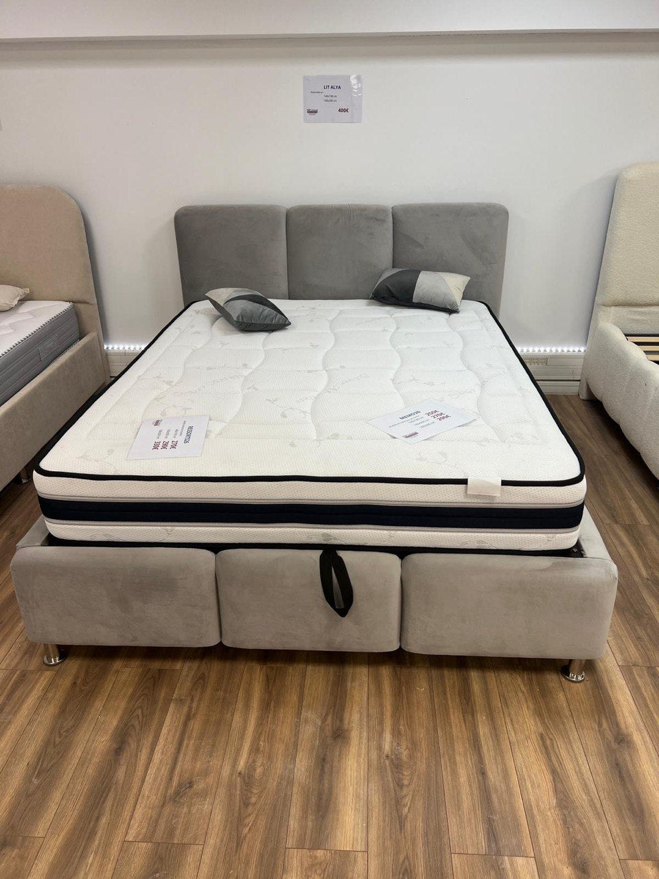 Lit coffre Alya en velours anti-tache gris clair, tête de lit à panneaux capitonnés, présenté avec un matelas en showroom